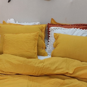 May include: A yellow linen duvet cover with a yellow, white, and orange pillow arrangement. The pillows have pom-pom trim.