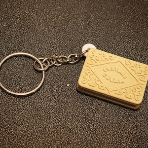 May include: A beige rectangular keychain with a biscuit design, featuring embossed details and text. The keychain has a silver metal ring and chain.