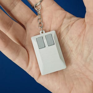 May include: A white remote control keyring with two grey buttons. The keyring has a silver metal ring and chain. The remote is held in a hand against a blue background.