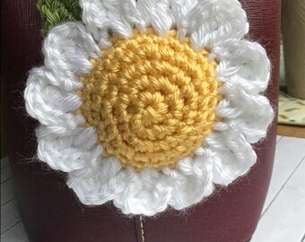 May include: A crochet daisy flower bag charm featuring a bright yellow center, white petals, and a single green leaf. The flower is attached to a metal chain and clasp, currently hanging from a dark burgundy leather handbag with visible stitching.