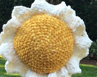 May include: A large, handmade crochet daisy pillow. The center is a textured golden yellow, surrounded by fluffy white petals. The pillow is round and appears soft, perfect for home decor.