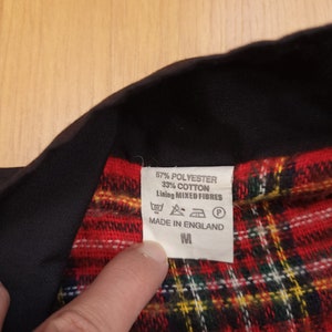 Peut inclure: Une chemise &agrave; carreaux noirs et rouges avec un col noir et une &eacute;tiquette blanche indiquant "67% Polyester 33% Coton Doublure Fibres Mixtes" et "Fabriqu&eacute; en Angleterre". L'&eacute;tiquette indique &eacute;galement la taille "M".