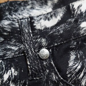 May include: Close-up of black denim jeans with a white abstract pattern. The jeans have a silver button with the text "100% RAW" and a small pocket. The fabric is textured and the stitching is visible.