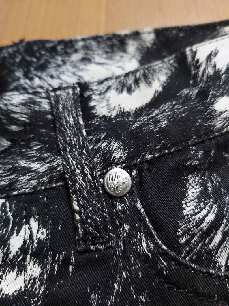 May include: Close-up of black denim jeans with a white abstract pattern. The jeans have a silver button with the text "100% RAW" and a small pocket. The fabric is textured and the stitching is visible.