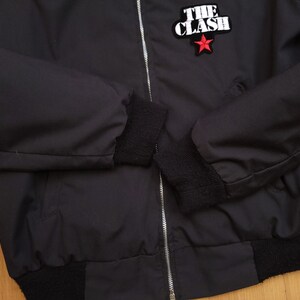 Peut inclure: Blouson noir avec fermeture &eacute;clair et poignets c&ocirc;tel&eacute;s. Le blouson pr&eacute;sente une &eacute;toile rouge et le texte "THE CLASH" en blanc.