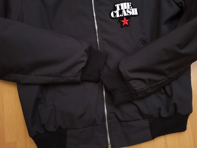 Peut inclure: Blouson noir avec fermeture &eacute;clair et poignets c&ocirc;tel&eacute;s. Le blouson pr&eacute;sente une &eacute;toile rouge et le texte "THE CLASH" en blanc.