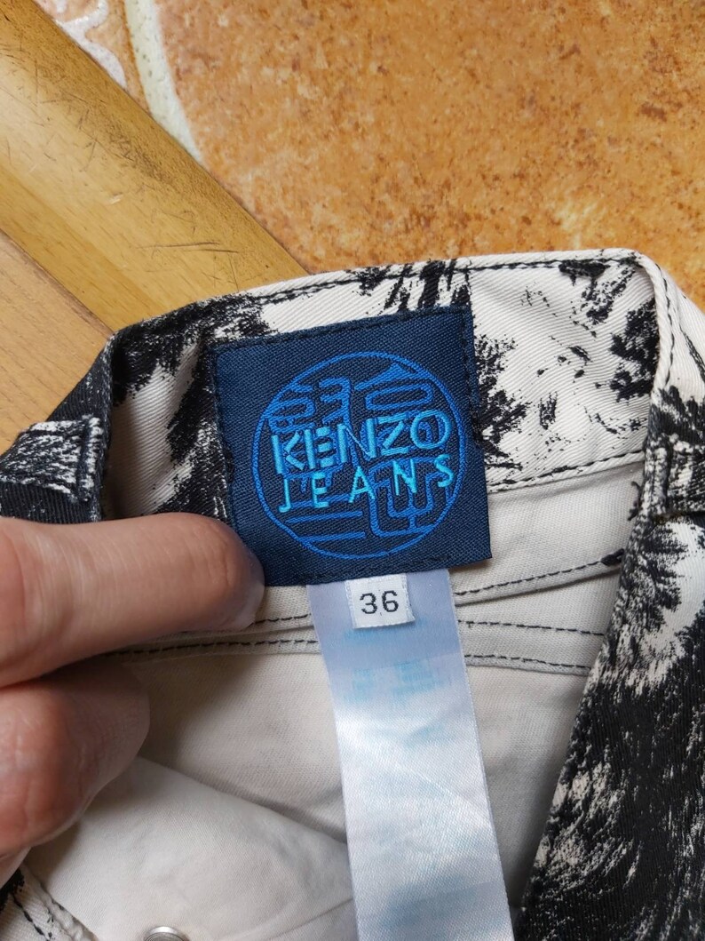 May include: Close-up of a pair of jeans with a black and white pattern. The blue label reads "KENZO JEANS" and the size tag shows "36". The jeans are made of a light-coloured fabric.