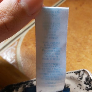 May include: A white clothing label with blue text, attached to a garment with a black and white pattern. The label includes care instructions, such as "wash at 40 degrees" and "no tumble dry."