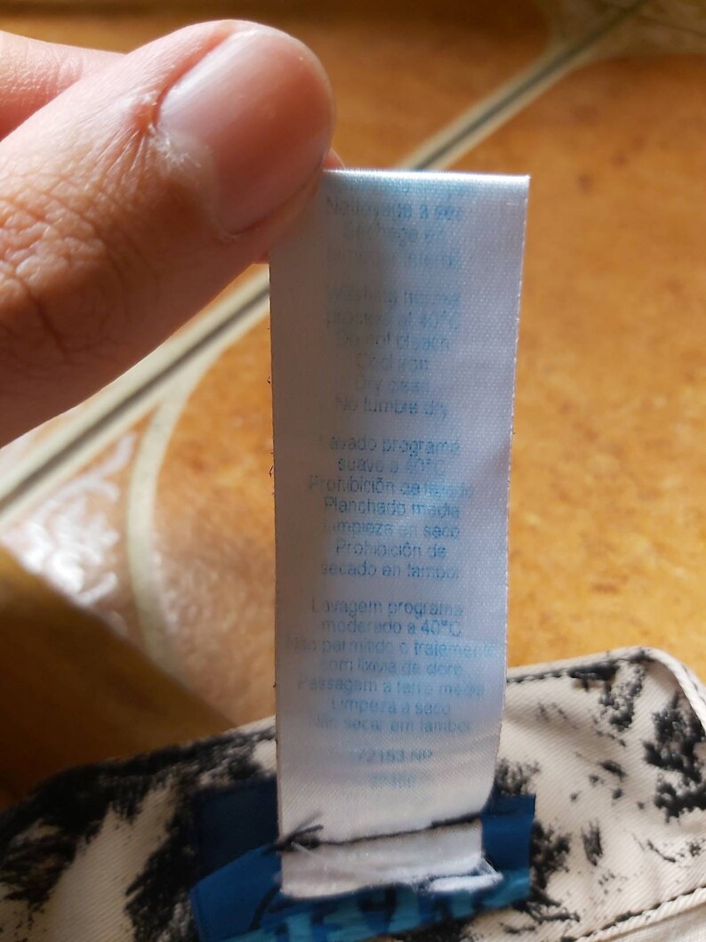 May include: A white clothing label with blue text, attached to a garment with a black and white pattern. The label includes care instructions, such as "wash at 40 degrees" and "no tumble dry."