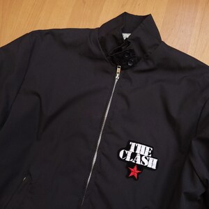 Peut inclure: Une veste noire zipp&eacute;e avec une &eacute;toile rouge et le texte "THE CLASH" brod&eacute; sur le devant.