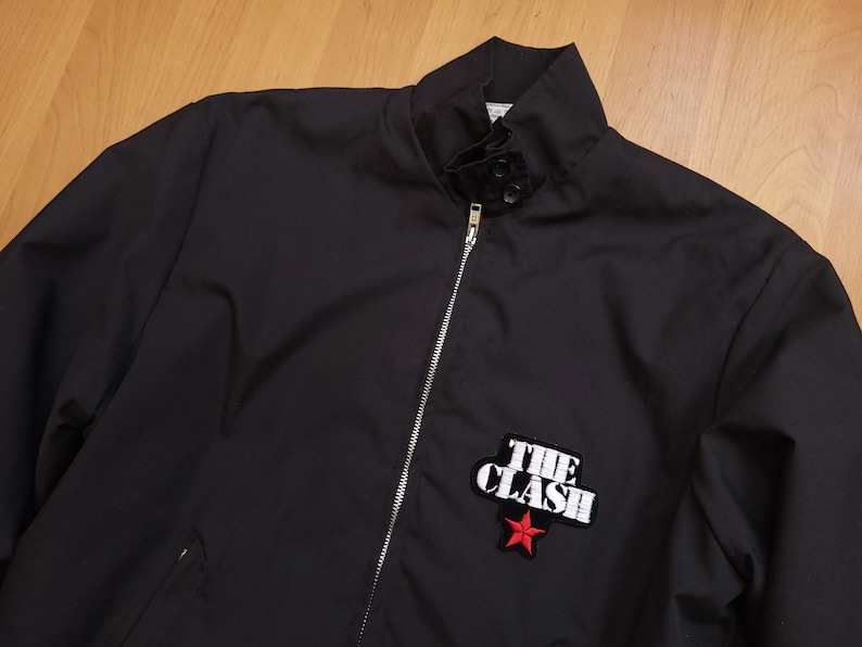 Peut inclure: Une veste noire zipp&eacute;e avec une &eacute;toile rouge et le texte "THE CLASH" brod&eacute; sur le devant.