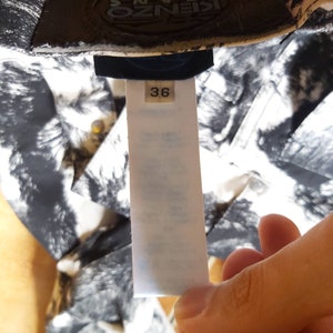 May include: A garment tag with the number 36, held by a hand. The background shows a black and white patterned fabric. The brand name "KENZO JEANS" is visible on the garment. The tag provides size information.