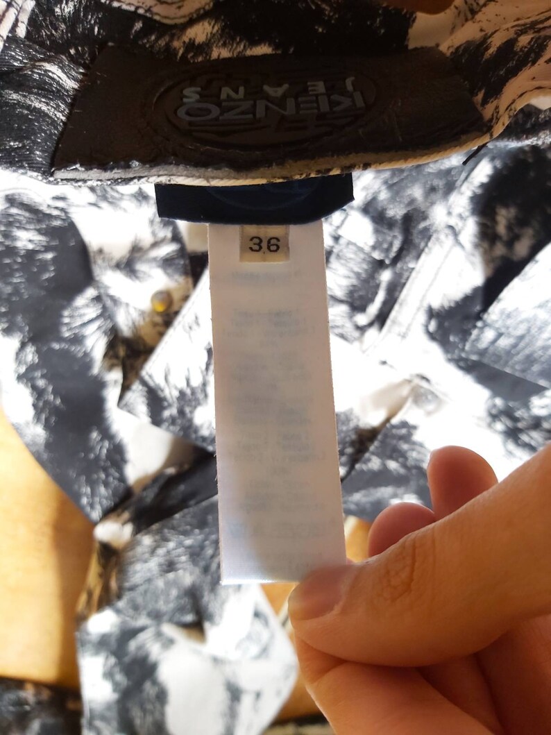 May include: A garment tag with the number 36, held by a hand. The background shows a black and white patterned fabric. The brand name "KENZO JEANS" is visible on the garment. The tag provides size information.
