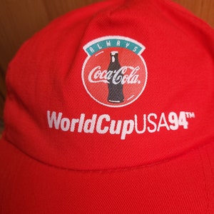 May include: Red baseball cap with a white Coca-Cola logo and the text "Always Coca-Cola World Cup USA 94" embroidered on the front.