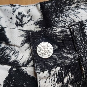 May include: Detail of jeans featuring a black and white abstract design. The jeans have a silver button with the word "JANSEN" around the edge. The fabric's pattern is a striking contrast of black and white, creating a distinctive look.