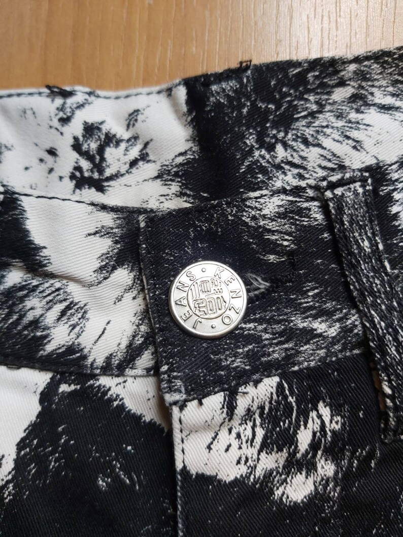 May include: Detail of jeans featuring a black and white abstract design. The jeans have a silver button with the word "JANSEN" around the edge. The fabric's pattern is a striking contrast of black and white, creating a distinctive look.