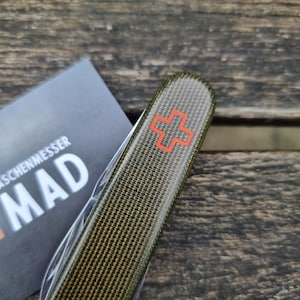 Puede incluir: Un mango de navaja multiusos con un acabado texturizado en color verde oliva similar a la tela y un emblema de cruz suiza naranja. El mango está colocado junto a una tarjeta negra con el texto parcial en blanco TASCHENMESSER MAD impreso.