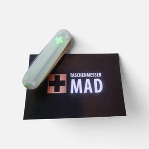 Op de afbeelding: Een zakmes met een doorschijnende, lichtgevende behuizing met een felgroen kruis-embleem, rustend op een zwarte rechthoekige kaart bedrukt met de tekst TASCHENMESSER MAD en een groot zwart kruispictogram.
