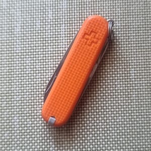Puede incluir: Una navaja de bolsillo naranja con un mango texturizado y un llavero plateado. La navaja presenta un emblema de cruz en el lateral. La hoja es parcialmente visible, lo que sugiere m&uacute;ltiples herramientas en su interior.