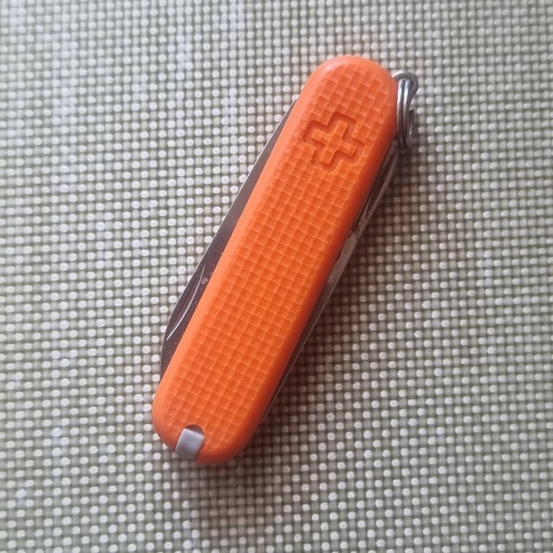 Puede incluir: Una navaja de bolsillo naranja con un mango texturizado y un llavero plateado. La navaja presenta un emblema de cruz en el lateral. La hoja es parcialmente visible, lo que sugiere m&uacute;ltiples herramientas en su interior.