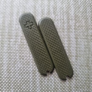 Puede incluir: Dos cachas de cuchillo de repuesto verde oliva con una superficie texturizada y un emblema de cruz. Las cachas son rectangulares con extremos redondeados y una peque&ntilde;a muesca en la parte inferior. Piezas de repuesto.