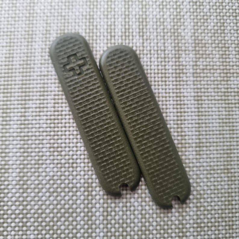 Puede incluir: Dos cachas de cuchillo de repuesto verde oliva con una superficie texturizada y un emblema de cruz. Las cachas son rectangulares con extremos redondeados y una peque&ntilde;a muesca en la parte inferior. Piezas de repuesto.