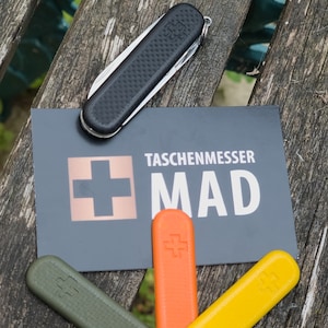 Puede incluir: Una navaja de bolsillo negra con un mango texturizado descansa sobre una tarjeta que dice "TASCHENMESSER MAD". Debajo de la tarjeta hay tres reemplazos de mango de cuchillo de colores: verde, naranja y amarillo. Los art&iacute;culos est&aacute;n sobre una superficie de madera desgastada.