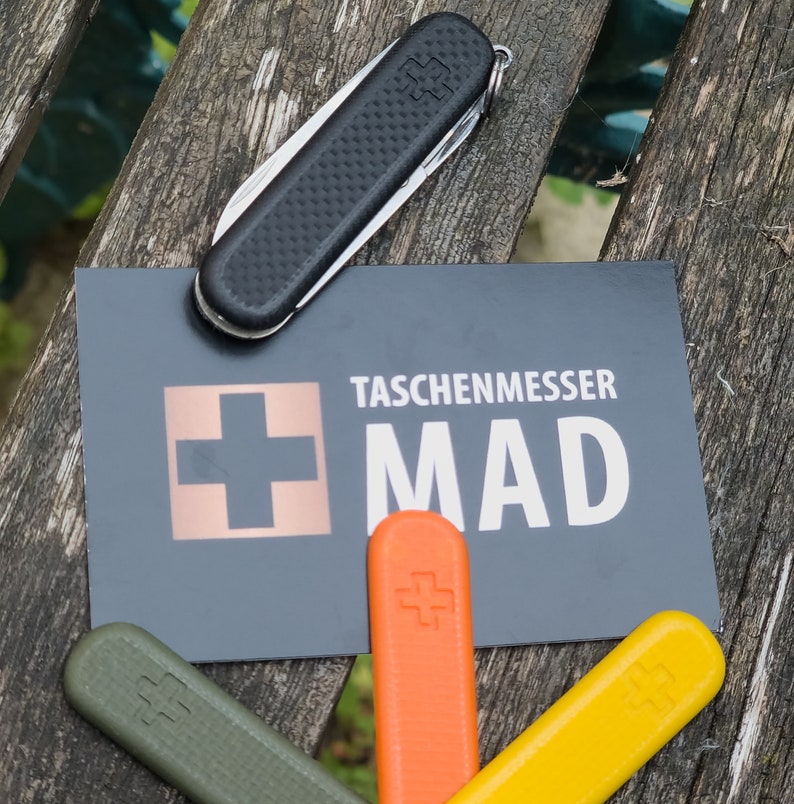 Puede incluir: Una navaja de bolsillo negra con un mango texturizado descansa sobre una tarjeta que dice "TASCHENMESSER MAD". Debajo de la tarjeta hay tres reemplazos de mango de cuchillo de colores: verde, naranja y amarillo. Los art&iacute;culos est&aacute;n sobre una superficie de madera desgastada.