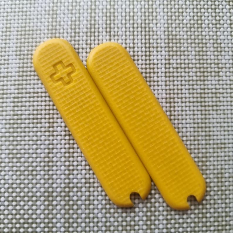 Puede incluir: Dos cachas de repuesto de color amarillo brillante para una navaja de bolsillo. Una cacha presenta un s&iacute;mbolo de cruz rebajado, y ambas tienen un patr&oacute;n texturizado en forma de rejilla. Las cachas tienen una parte superior curvada y una peque&ntilde;a muesca en la parte inferior.