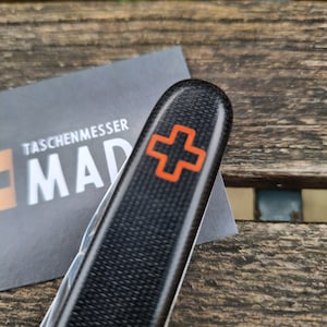 May include: A black pocket knife with a carbon fibre texture and an orange cross emblem. The knife is partially open, revealing a silver blade. A business card with the text "TASCHENMESSER MAD" is in the background.