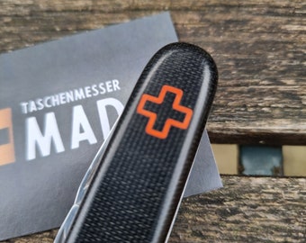 May include: A black pocket knife with a carbon fibre texture and an orange cross emblem. The knife is partially open, revealing a silver blade. A business card with the text "TASCHENMESSER MAD" is in the background.