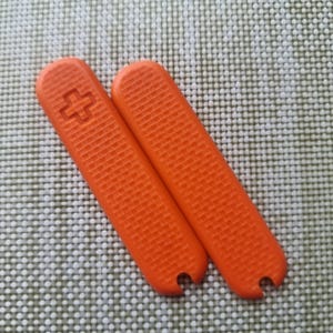 Puede incluir: Dos cachas de cuchillo de repuesto de color naranja con una superficie texturizada. Una de las cachas tiene un s&iacute;mbolo de cruz rebajado. Las cachas son rectangulares con la parte superior curvada y una peque&ntilde;a muesca en la parte inferior.
