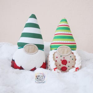 May include: Two plush gnome figures. The left gnome has a green and white striped hat, a white beard, and a red and black plaid base. The right gnome has a green, white, and red striped hat, two white braided pigtails, and a tan body with red and green polka dots.