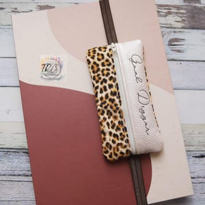 May include: A pencil case with a leopard print pattern and the text "Goal Digger" in cursive. The case is white with a zipper closure and is resting on a pink and burgundy notebook.