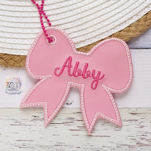 May include: A light pink bow-shaped tag featuring the name Abby embroidered in a darker pink script font. The tag includes a metal grommet at the top attached to a bright pink ball chain, with visible stitching along the edges of the bow shape.
