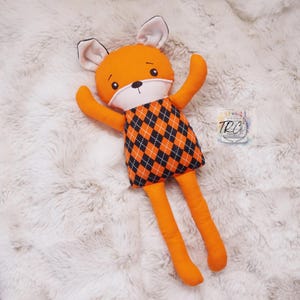 May include: A plush fox toy with bright orange limbs and ears, featuring a white face with embroidered black eyes and nose. The fox wears a black and orange diamond-patterned tunic. A small square label with the text TRC is visible nearby.