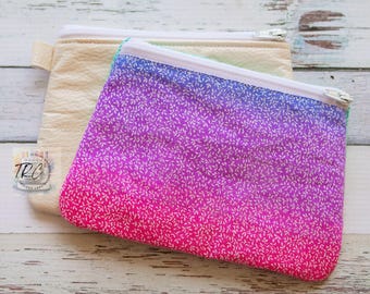 May include: Two zippered pouches, one is a plain beige fabric and the other is a pink to purple ombre fabric with a white floral pattern.