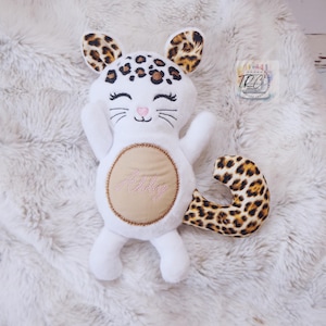 May include: A white plush cat toy featuring leopard print ears and a matching curled tail. The face has embroidered closed eyes, whiskers, and a pink nose. A tan circular patch on the belly displays the embroidered name Lily in pink script.