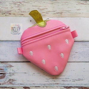 Puede incluir: Una bolsa rosa con cremallera en forma de fresa con una parte superior de purpurina dorada. La bolsa tiene detalles bordados blancos que se asemejan a semillas.