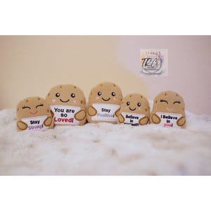 Op de afbeelding: Vijf beige pluche aardappelfiguren met geborduurde gezichten en armen die witte bordjes vasthouden. De bordjes bevatten de tekst: Stay Strong!, You are so Loved!, Stay Positive!, Believe In yourself! en I Believe In you! met kleurrijke accenten.