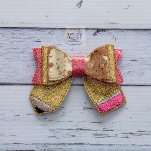 Pode incluir: Um laço de glitter rosa e dourado com um design de lápis. O laço é feito de tecido e tem uma textura de cortiça.