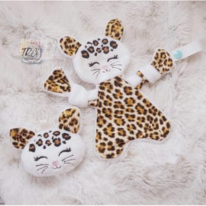 May include: A plush leopard-themed security blanket and a matching small plush head. Both items feature white fabric with brown and black leopard print accents, embroidered closed eyes, whiskers, and a pink nose. A white strap with a blue snap is attached.