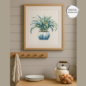 May include: A framed watercolor print of a potted plant with long green leaves and a blue and white patterned pot. The artwork is displayed on a light gray wall. A wooden peg rack, a striped towel, and kitchenware are in the foreground.