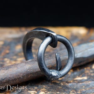 Puede incluir: Primer plano de un anillo artesanal de metal oscuro con un diseño único e entrelazado. El anillo presenta una banda circular conectada a un elemento curvo. El anillo descansa sobre una superficie metálica texturizada. El nombre de la tienda Etsy "Simas Designs" es visible.