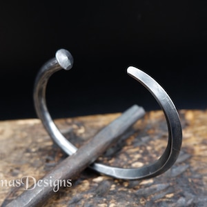 Op de afbeelding: Een donkergrijze metalen armband in de vorm van een gebogen spijker met een afgeronde kop aan het ene uiteinde en een taps toelopend punt aan het andere. De armband heeft een gladde, matte afwerking en ligt op een metalen staaf. Tekst Simas Designs.