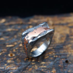 Op de afbeelding: Een zilveren ring met een getextureerd, asymmetrisch ontwerp. De ring heeft een koperkleurige inleg die contrasteert met het zilver. De ring wordt getoond op een houten oppervlak.
