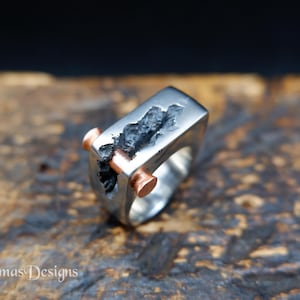 Op de afbeelding: Een zilverkleurige ring met een dikke, rechthoekige band met een centrale, grillige, donkere, getextureerde spleet. Een gladde, cilindrische, koperkleurige pin loopt horizontaal door het midden van de donkere, ruwe opening in het metaal.
