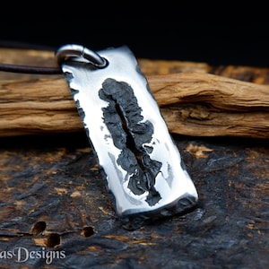 Op de afbeelding: Een rechthoekige zilveren hanger met een donker, getextureerd midden, hangend aan een bruin leren koord. De hanger heeft een robuuste, handgemaakte uitstraling. De merknaam "Simas Designs" is linksonder zichtbaar. Het is waarschijnlijk een sieraad.