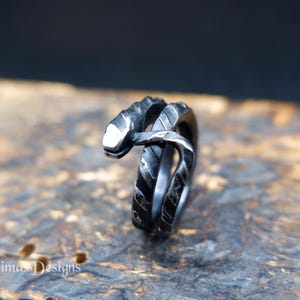 Könnte beinhalten: Ein dunkelgrauer Metallring in Form einer gewundenen Schlange mit einer texturierten, unebenen Oberfläche. Der Schlangenkopf befindet sich oben am Ringband, und der Körper windet sich um die Ringstruktur. Der Text Simas Designs ist sichtbar.