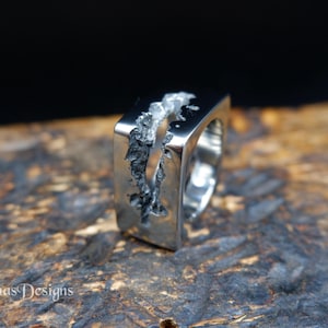 May include: A silver-toned metal ring featuring a smooth, polished rectangular face with a deep, jagged, and textured vertical fissure running through the center. The brand name Simas Designs is visible in the lower left corner of the image.
